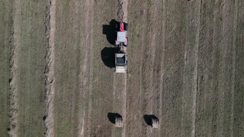 Tractor harvesting grass and making hay bales in field, aerial drone view. Aerial drone footage of a tractor working in an agricultural field during summer. The machine harvests grass and packs it 