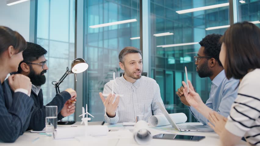 Caucasian male engineer explaining wind turbine model during team discussion with diverse colleagues at conference table. Professionals brainstorming renewable energy project in bright office. - Powered by Shutterstock - Get 15% off with code: PIKWIZARD15