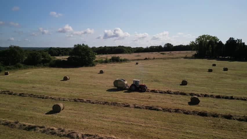 Tractor harvesting grass and making hay bales in field, aerial drone view. Aerial drone footage of a tractor working in an agricultural field during summer. The machine harvests grass and packs it 