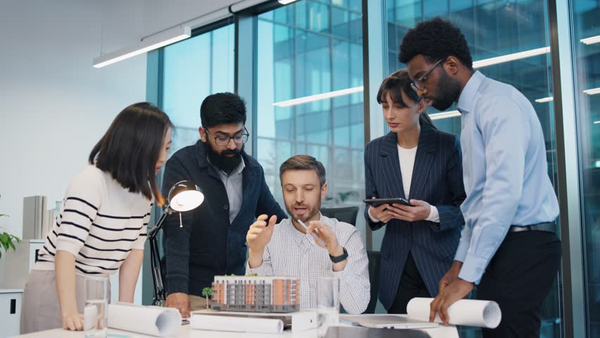 Caucasian man explaining concept while team of five professionals examining architectural model. Diverse colleagues standing around table in modern office, collaborating on building design project. - Powered by Shutterstock - Get 15% off with code: PIKWIZARD15