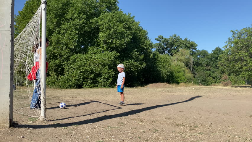 A young boy is focused on kicking a soccer ball on a dusty outdoor field, while a woman stands by the goal net. They enjoy a leisurely and active day under the clear blue sky, surrounded by green tree
