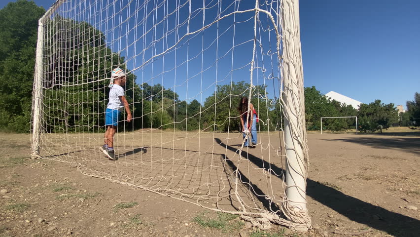 A child acts as a goalkeeper, standing playfully in a soccer net on a sunny dirt field, while a woman observes from a distance. The scene captures a lighthearted outdoor game amidst green trees.