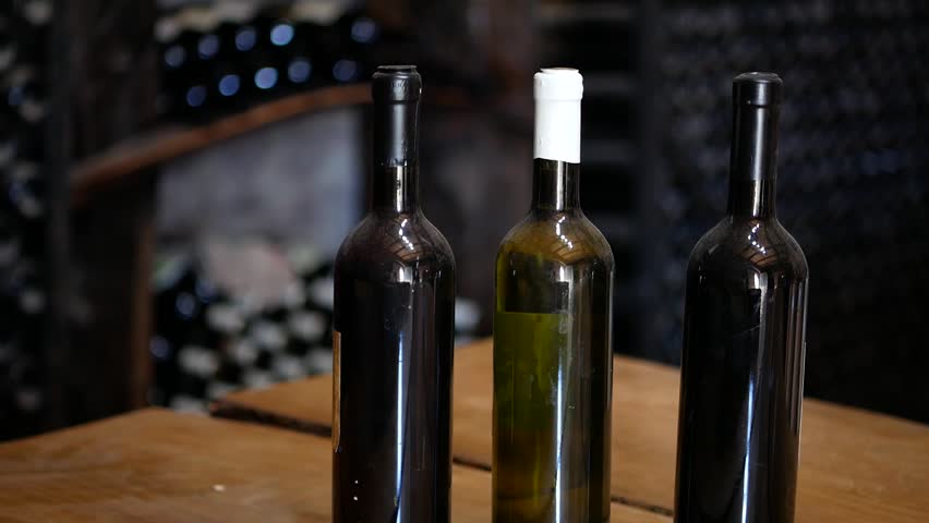 Bottles of wine in the wine cellar. Three bottles of three different wines are sitting on a barrel in the wine cellar. The bottles have no labels. Behind them are shelves with other wine bottles