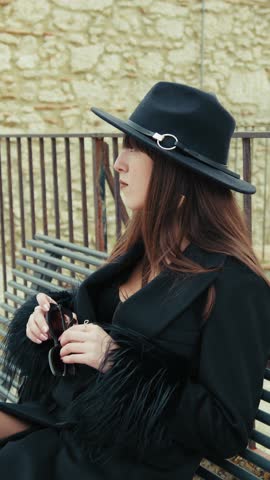 Elegant Woman In Black Coat Sitting On A Bench In Her Town And Looking Around 