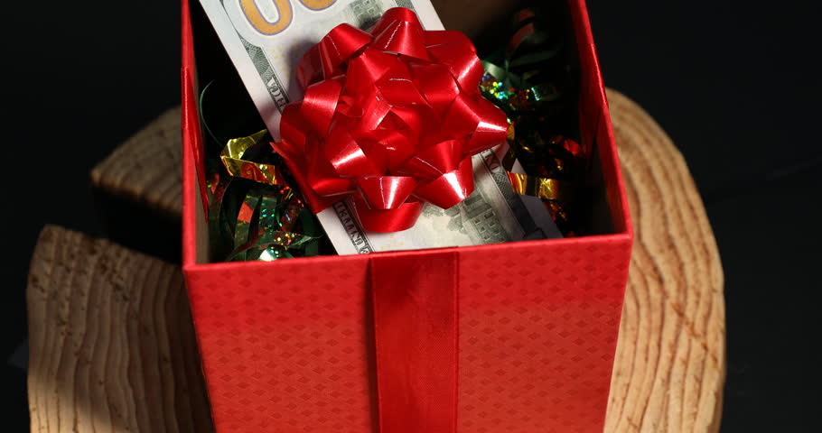 Gift box with dollar banknotes and tinsel on wooden stump against black background, closeup