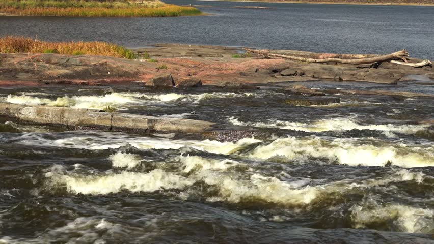 River rapids with fast flowing water and white foam. Waves crashing over rocks with rocky shoreline in the background. Scenic wilderness landscape with powerful current.