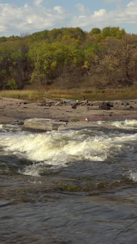 Fast river rapids flowing over rocks with autumn forest in the background. The scene shows strong white water movement, rocky shoreline, and seasonal trees under a partly cloudy sky.