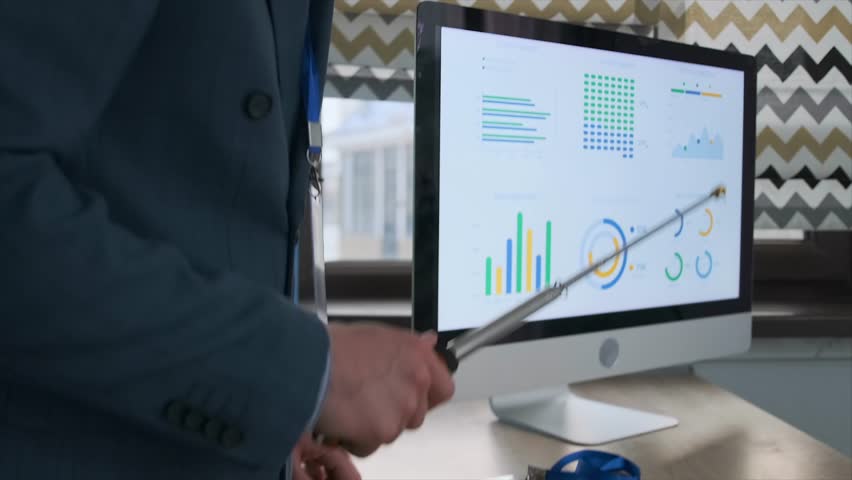 Close-up shot of a young businessman in a suit presenting data with a metal pointer. His corporate badge is visible, and screen displays business graphs and analytics. Professional data presentation.
