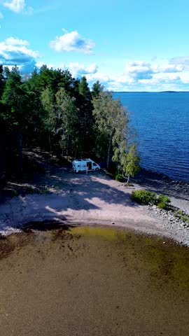 A couple of men and women stroll along the sandy shore of Lake Saimaa Finland, surrounded by lush greenery and a tranquil atmosphere. A camper van is parked on the beach
