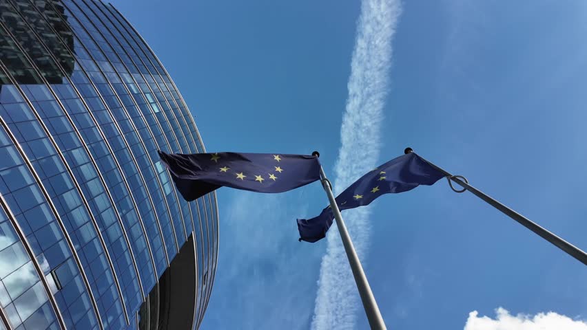 EU flags flutter in front of the Lex Building in Brussels with airplane contrail across the clear blue sky.