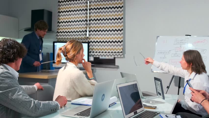 Man in a formal suit giving a business presentation in front of a team in a modern office. Charts and diagrams are displayed on the monitor. Whiteboard shows business planning steps.