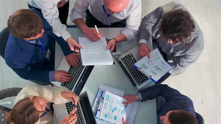 Top view of a business team working around a table with laptops and documents. A leader is signing a contract while others express different opinions and gestures. Concept of corporate negotiation.