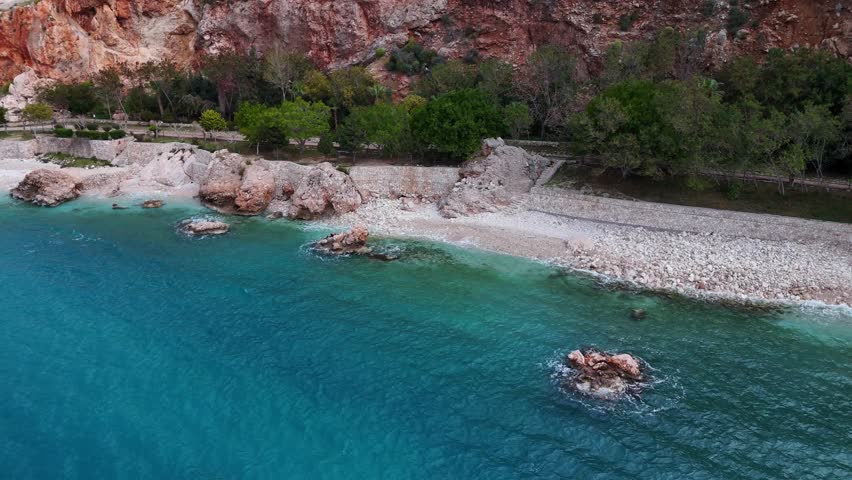 Aerial view of the seaside promenade and walking path along the coast in Kalkan, Turkey. Red cliffs, green hills, and turquoise sea form a picturesque Mediterranean landscape.
