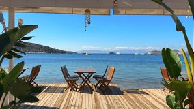 Seaside dining deck with wooden tables and chairs overlooking a beautiful turquoise bay, sailboats, and distant hills under a clear blue sky. - Powered by Shutterstock - Get 15% off with code: PIKWIZARD15