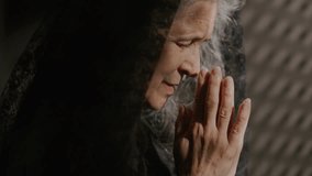 Senior woman in black lace veil praying deeply to God with her eyes closed and head bowed to clasped hands in confessional booth. Close-up profile view - Powered by Shutterstock - Get 15% off with code: PIKWIZARD15