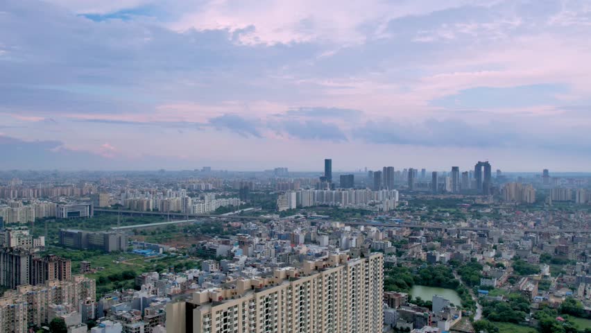 aerial drone hyperlapse flying backwards showing the multi-storey buildings in delhi, gurgaon surrounded by green fields and small houses with giant fluffy monsoon clouds flying overhead