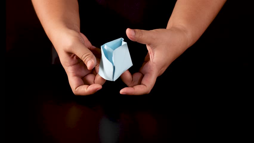 Person folds blue paper into origami boat, overhead view, dramatic lighting, dark background, steady camera