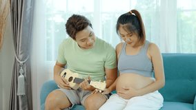 Beautiful pregnant asian woman and young man are smiling and playing Ukulele or acoustic guitar on sofa, Husband playing Ukulele for pregnant woman and baby in the womb, concept relax day and holiday - Powered by Shutterstock - Get 15% off with code: PIKWIZARD15