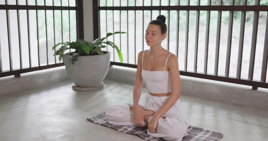 Young woman in white outfit tries to meditate but gets easily distracted, looks around, checks her phone, and quits practice. Conceptual footage for distraction, stress, technology, and lifestyle.