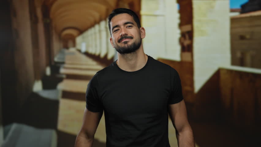 Young hispanic man posing in old university corridor with sunlight casting long shadows creating a contemplative and historical atmosphere.