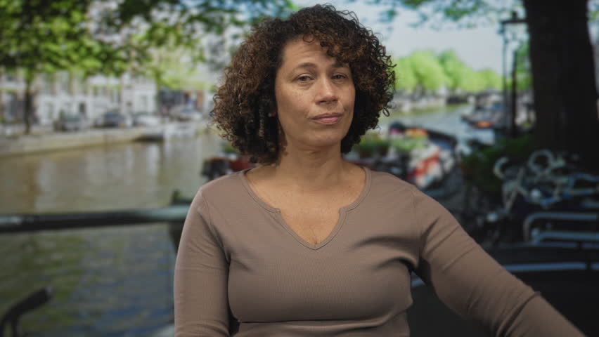 Woman finger pointing to watch on her wrist while standing on a canal street lined with bicycles and water; impatience.