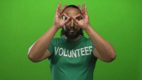 Man making binoculars with hands wearing volunteer tshirt against green backdrop in studio; playful outreach. - Powered by Shutterstock - Get 15% off with code: PIKWIZARD15