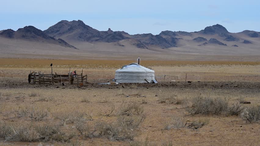 In a remote and vast Mongolian landscape, a traditional yurt (ger) and a wooden animal pen stand before a rugged mountain range, capturing a nomadic herder's lifestyle.