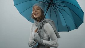 Woman wearing knit hat and scarf holding blue umbrella, smiling and looking up in gray studio backdrop with winter gloves visible; optimism. - Powered by Shutterstock - Get 15% off with code: PIKWIZARD15