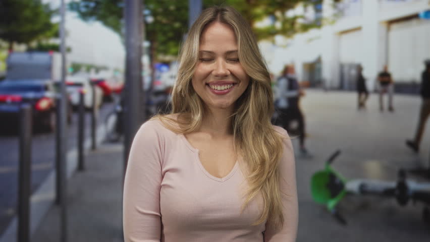 Woman with blonde hair smiles openly with shining eyes on city sidewalk; happiness confidence optimism warmth.