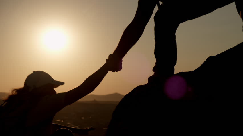 Silhouette of man hiker extending helping hand to friend while climbing mountain