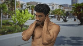 Shirtless young hispanic man holds neck with both hands on a sunny city street near palm trees and tall buildings; stress tension discomfort anxiety. - Powered by Shutterstock - Get 15% off with code: PIKWIZARD15
