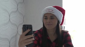Cheerful young woman wearing a Santa hat makes a video call on her smartphone at home. Cozy holiday atmosphere with warm lighting, festive greetings and joyful emotions.  - Powered by Shutterstock - Get 15% off with code: PIKWIZARD15