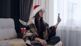 Smiling young woman in a Santa hat sits on a cozy sofa wrapped in a blanket, waving at the phone camera while talking on a video call during Christmas holidays.  - Powered by Shutterstock - Get 15% off with code: PIKWIZARD15