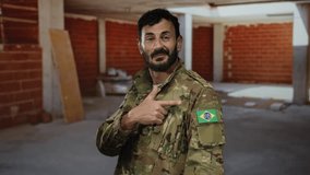 Hispanic man in military uniform with brazilian flag in a construction site, pointing at his shoulder, surrounded by unfinished brick walls, exuding confidence and focus. - Powered by Shutterstock - Get 15% off with code: PIKWIZARD15