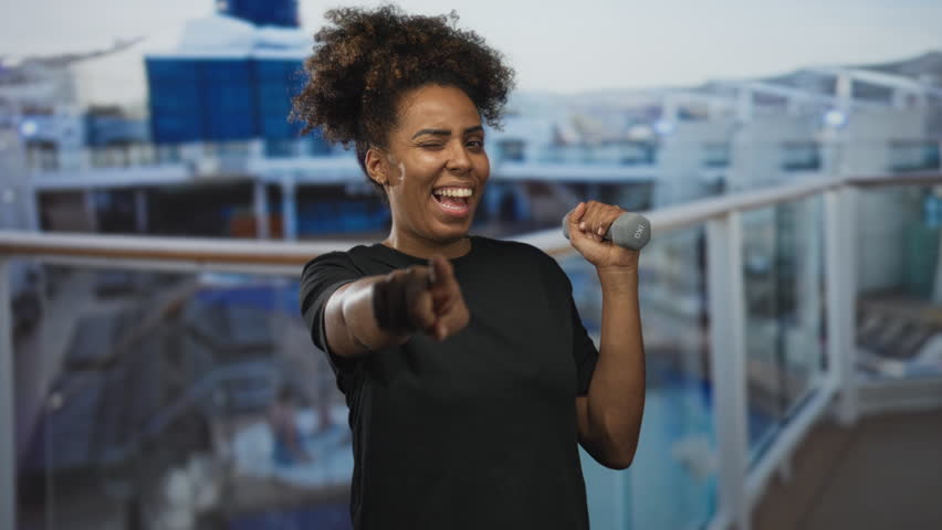 Woman points finger while winking and holding a dumbbell on a cruise ship deck building during an outdoor workout session; confidence motivation.
