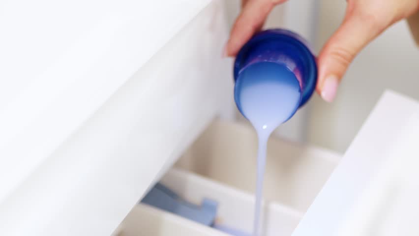 Hand pouring liquid laundry detergent or fabric softener into washing machine — clothes washing and home cleaning routine concept
