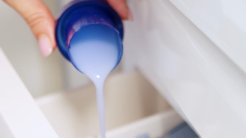 Hand pouring liquid laundry detergent or fabric softener into washing machine — clothes washing and home cleaning routine concept