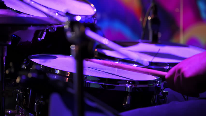 Live concert performance, a drummer energetically playing on stage. The scene captures the dynamic motion of drumsticks hitting cymbals and drums