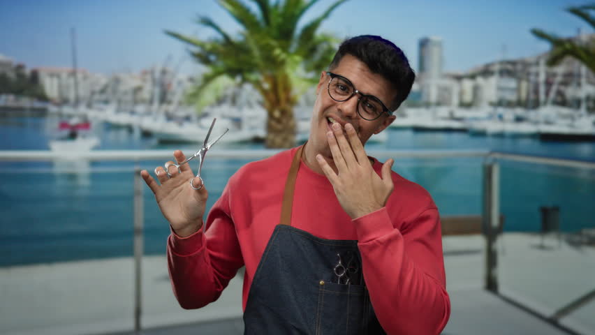Young man with scissors in a seaside port setting wearing glasses and apron showcasing a playful expression against a backdrop of boats and palm trees.