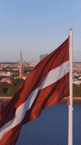 Aerial view of Riga, Latvia featuring the Latvian flag fluttering, the Daugava River, Vansu Bridge, and parts of the city's skyline with motion.