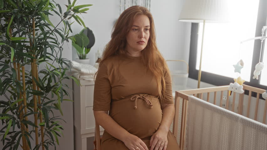 Pregnant caucasian woman cradles belly and supports lower back in bright nursery near wooden crib; anticipation.