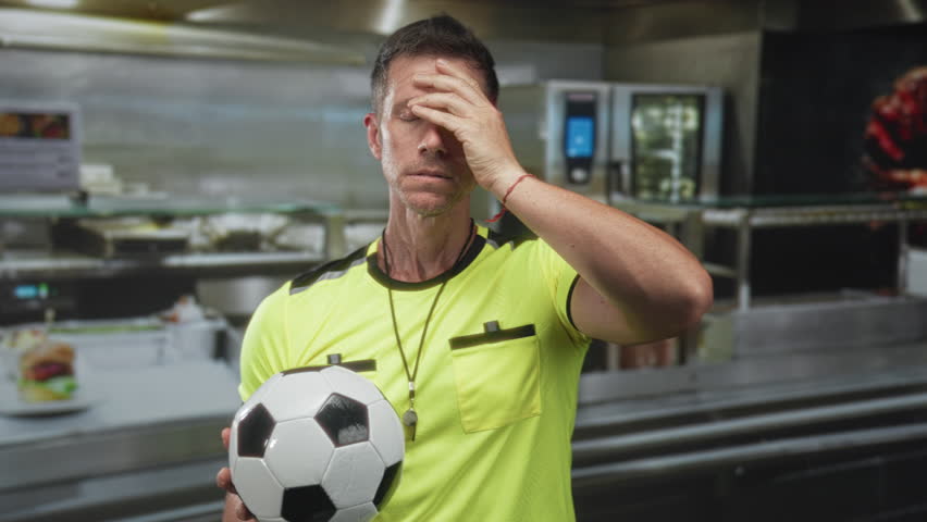 Man referee holding soccer ball, hand to forehead for headache in kitchen building; fatigue perseverance.