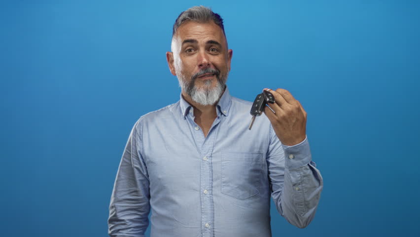 Man smiling and holding car keys while extending his hand for a handshake in blue studio; trust sale friendly.