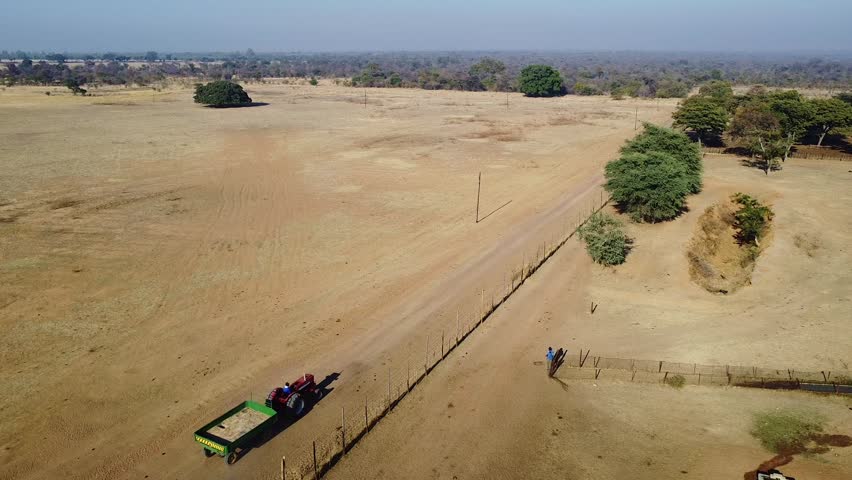 This aerial shot captures a vehicle on a dirt road through a rural landscape. The scene showcases the beauty of the countryside, with trees and open fields.