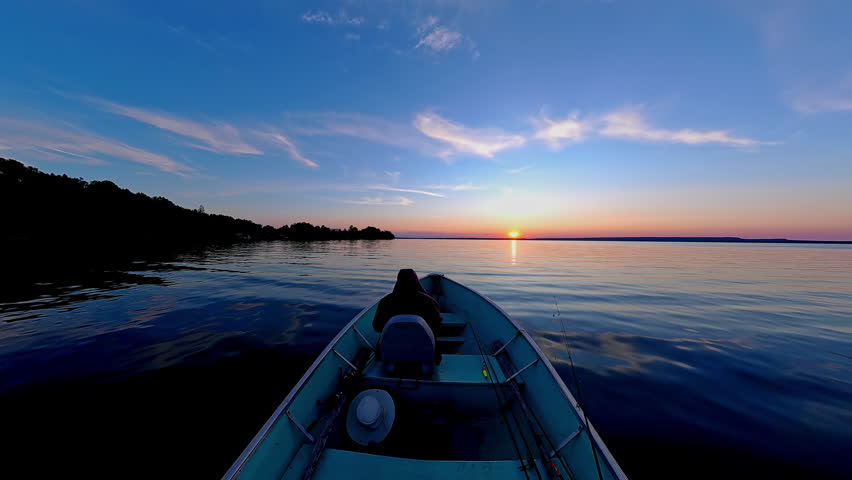 Boat with anglers tourists gliding during golden hour sunset with land skyline silhouetted. Best time for fishing game. View of sun set horizon at lake from boat. Cinematic view of evening leisure.
