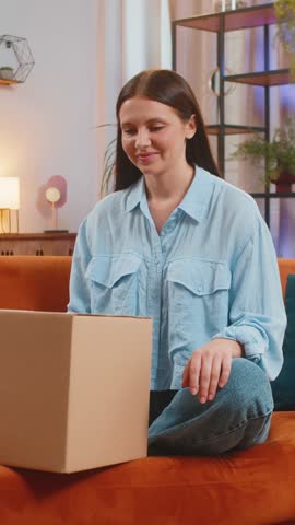 Happy Caucasian young woman unpacking delivery parcel sitting on home couch. Girl shopper online shop customer opening cardboard box receiving purchase gift by fast postal shipping at room apartment.