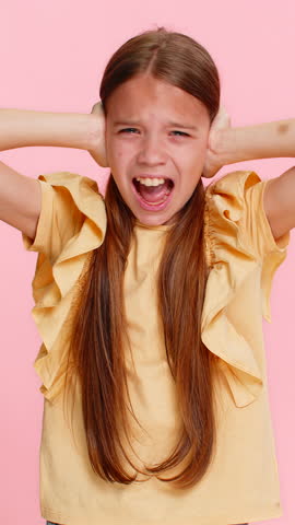 Young child girl covers ears with both hands, opening eyes wide in sudden fear panic caused by a loud sound or noise. School kid isolated on pink background, reacting emotionally with dramatic gesture