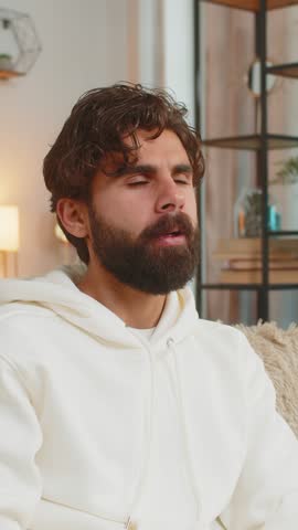 Upset Indian young man in hoodie holds hand over sore throat. Painful neck and frowning, thyroid disorders, suffering sore throat, tonsils inflammation. Hispanic guy male sitting on sofa at home room.