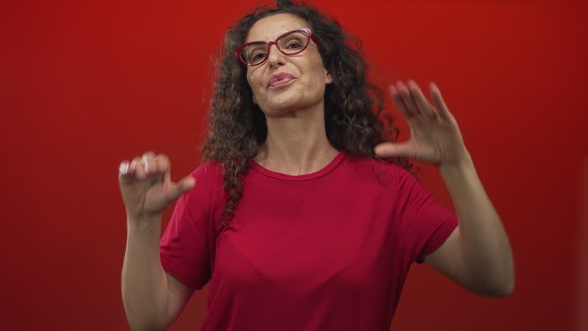 Hispanic woman wearing red glasses and red shirt framing smiling face with hands in studio red backdrop; playful joy spontaneity warmth.