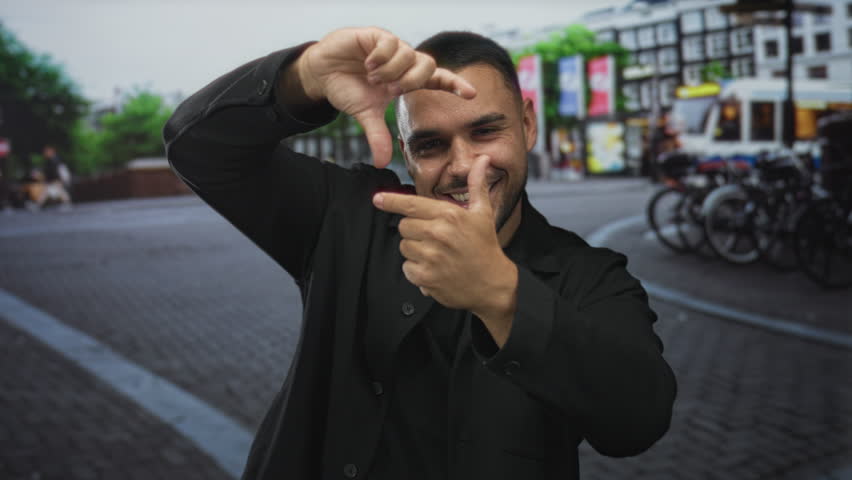 Man in black jacket smiling broadly with cheerful grin and framing scene with hands on urban street; creative joy.